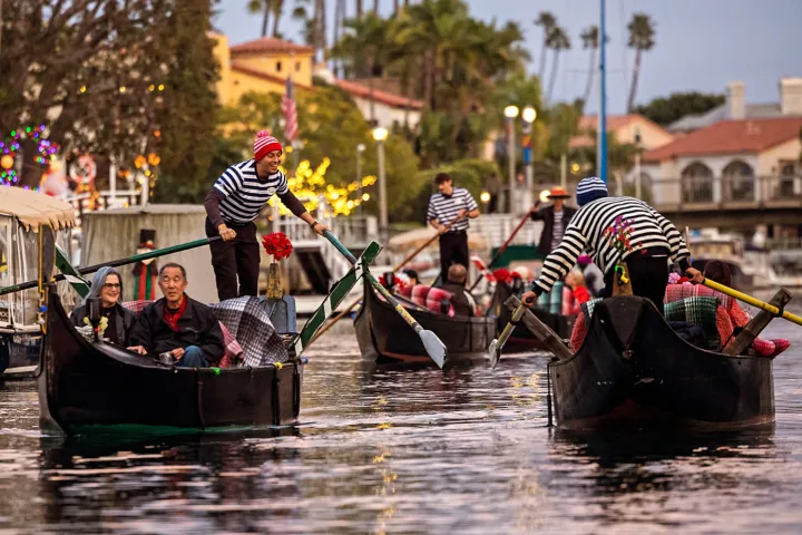 Gondoliers rowing gondolas with passengers on a scenic canal at sunset, surrounded by festive lights and palm trees.