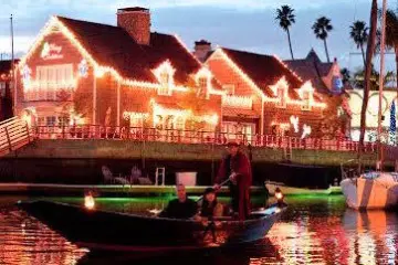 Boat with people on a canal at night, festive lights on houses.
